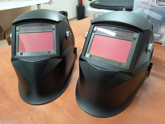 Two new black welding helmets sit side by side on a wooden workbench. Both have large rectangular red-tinted viewing windows and matte black plastic shells, appearing clean and unused. The helmets are identical in shape and size, positioned facing forward. In the background, workshop items like plastic storage boxes and tools are slightly visible, indicating a makerspace or workshop environment.