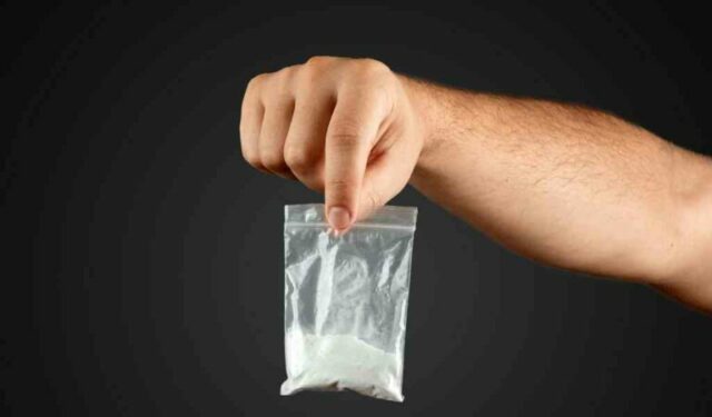 Male hand holds a plastic bag with cocaine on a blurred background, closeup. Social problem, drug addiction, death, addiction. Copy space.