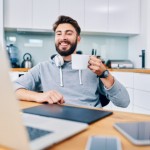 Joyful young web developer drinking coffee while taking break from work in home office