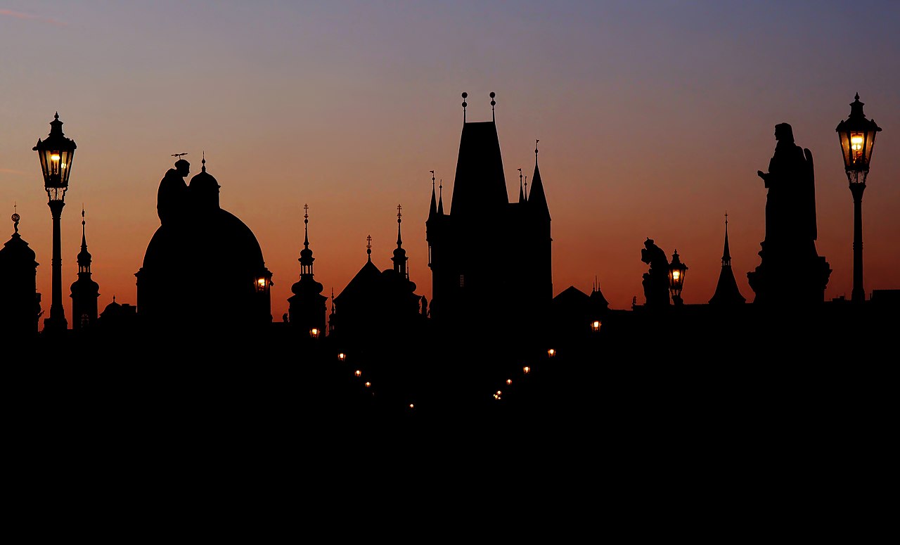 Dawn silhouette of Charles Bridge in Prague: gothic towers, baroque domes and bridge statues against an orange sky