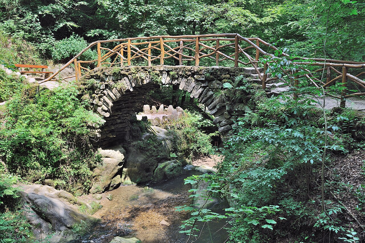森林中一座石拱桥横跨溪流，桥身覆苔藓，木质栏杆，周围绿植茂密，左侧步道有橙色长椅，下方溪流流经岩石。