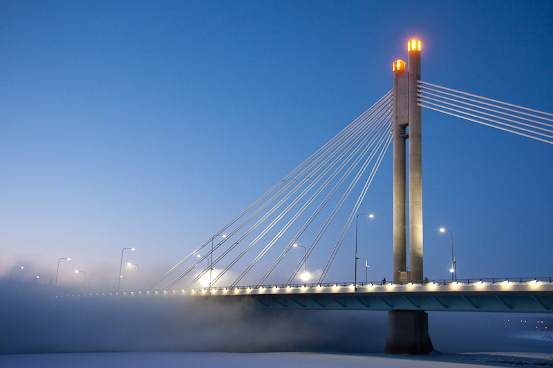 Winter night view of Rovaniemi's 'Lumberjack's Candle' cable-stayed bridge lit up, with mist over the river