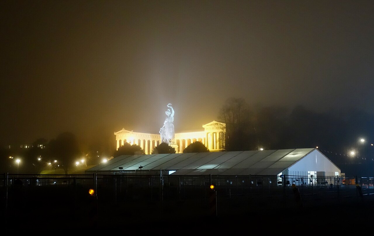 夜晚的Theresienwiese场地，有一座大型白色帐篷、围栏，背景是灯光照亮的巴伐利亚雕像和柱廊建筑，雾气弥漫，周边有路灯。