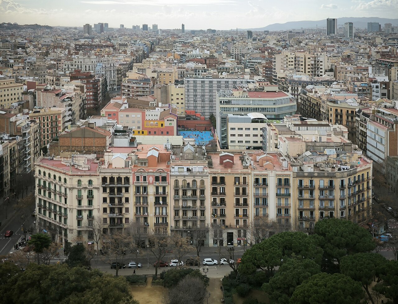 Aerial view over Barcelona's dense Eixample residential blocks with the city skyline and hills beyond