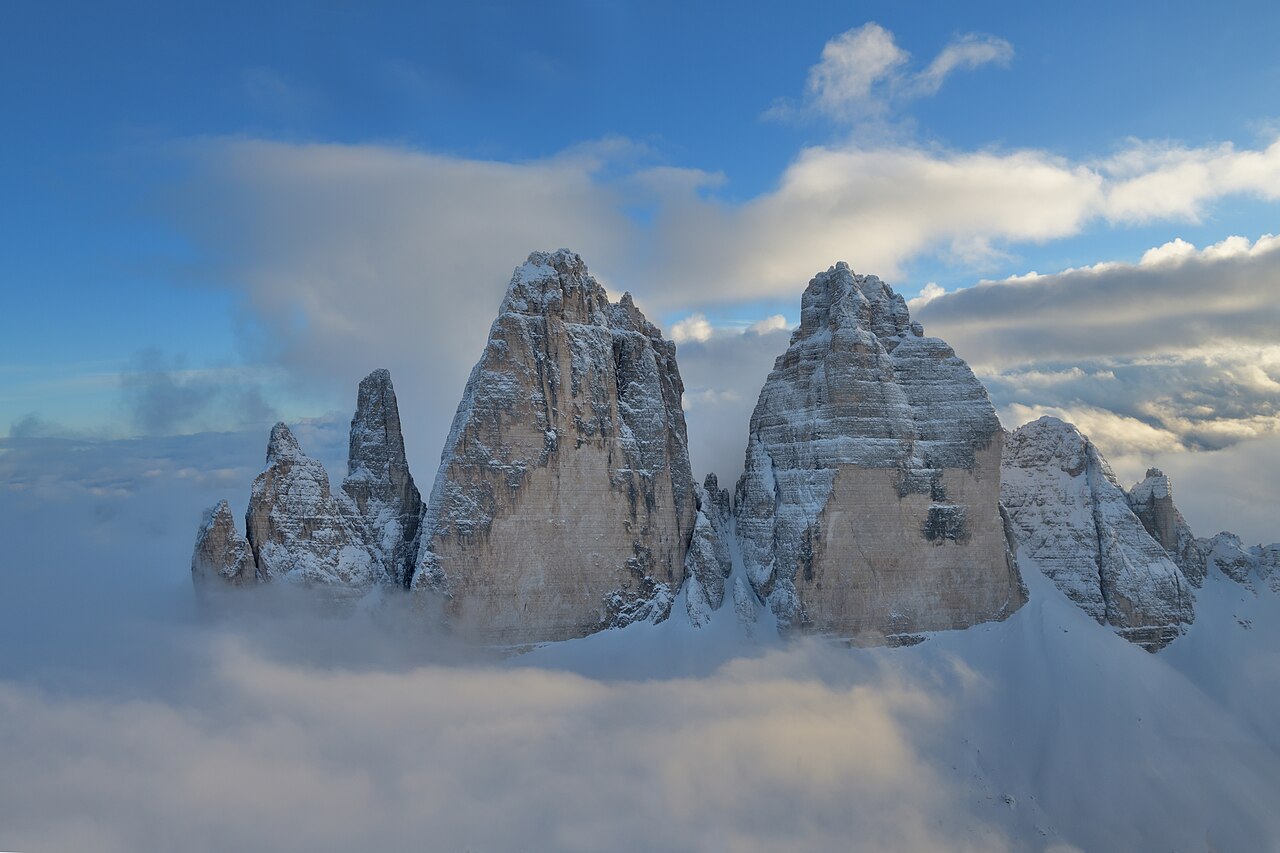 Tre Cime di Lavaredo的岩石山峰，部分覆盖积雪，被云雾和蓝天环绕。