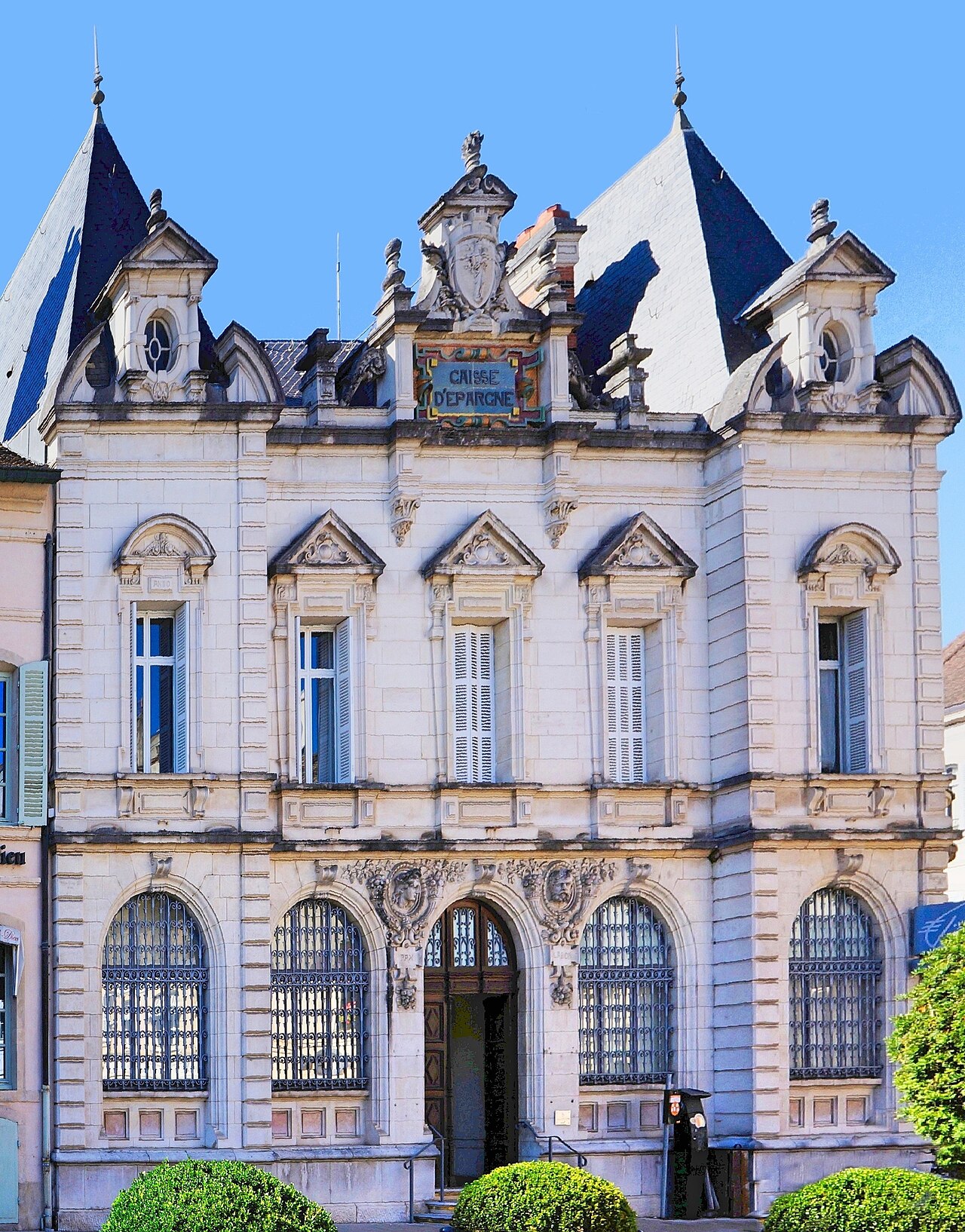 法国博讷的一栋浅色石质建筑，顶部有“Caisse d’Epargne”标识，底层拱窗带铁栅栏，门前有绿篱。
