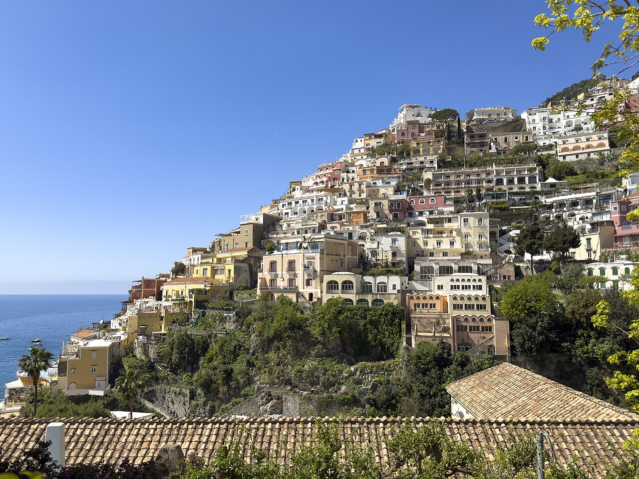 波西塔诺陡峭山坡上的彩色房屋群,下方是地中海,前景有红瓦屋顶和树木,蓝天晴朗。