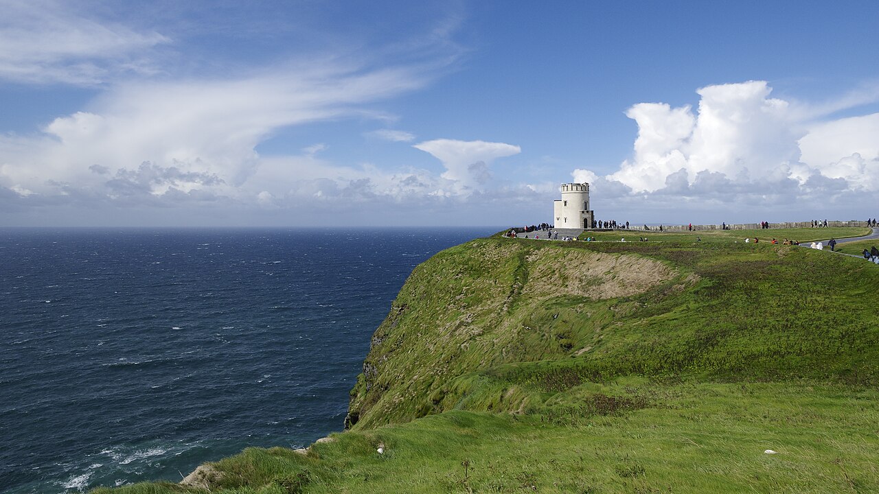 Green clifftop meadow at the Cliffs of Moher with visitors walking along the railing, O'Brien's Tower in the distance