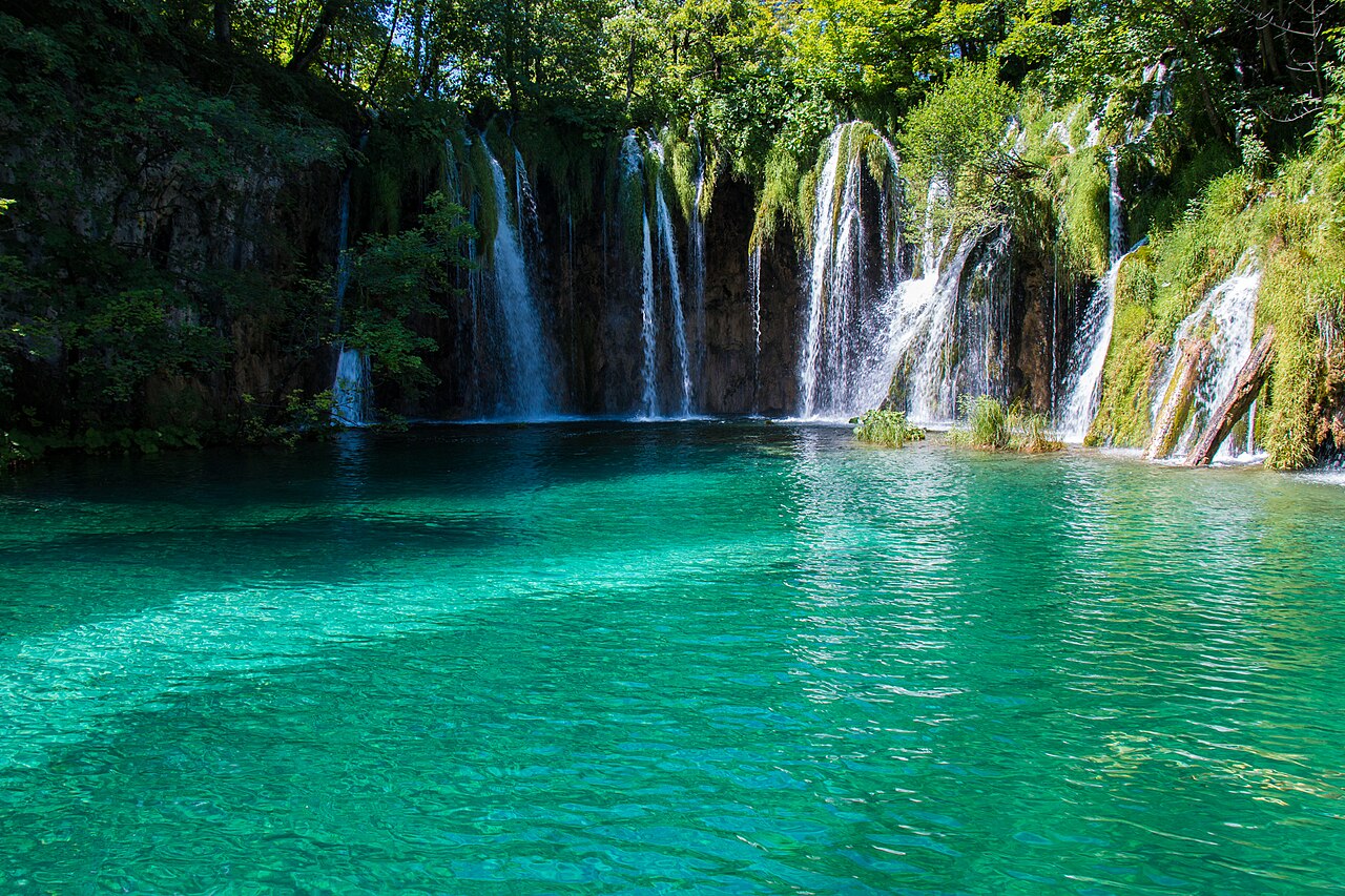 Emerald green lake surface of Plitvice Lakes, with multi-tiered limestone travertine waterfalls cascading through the forest