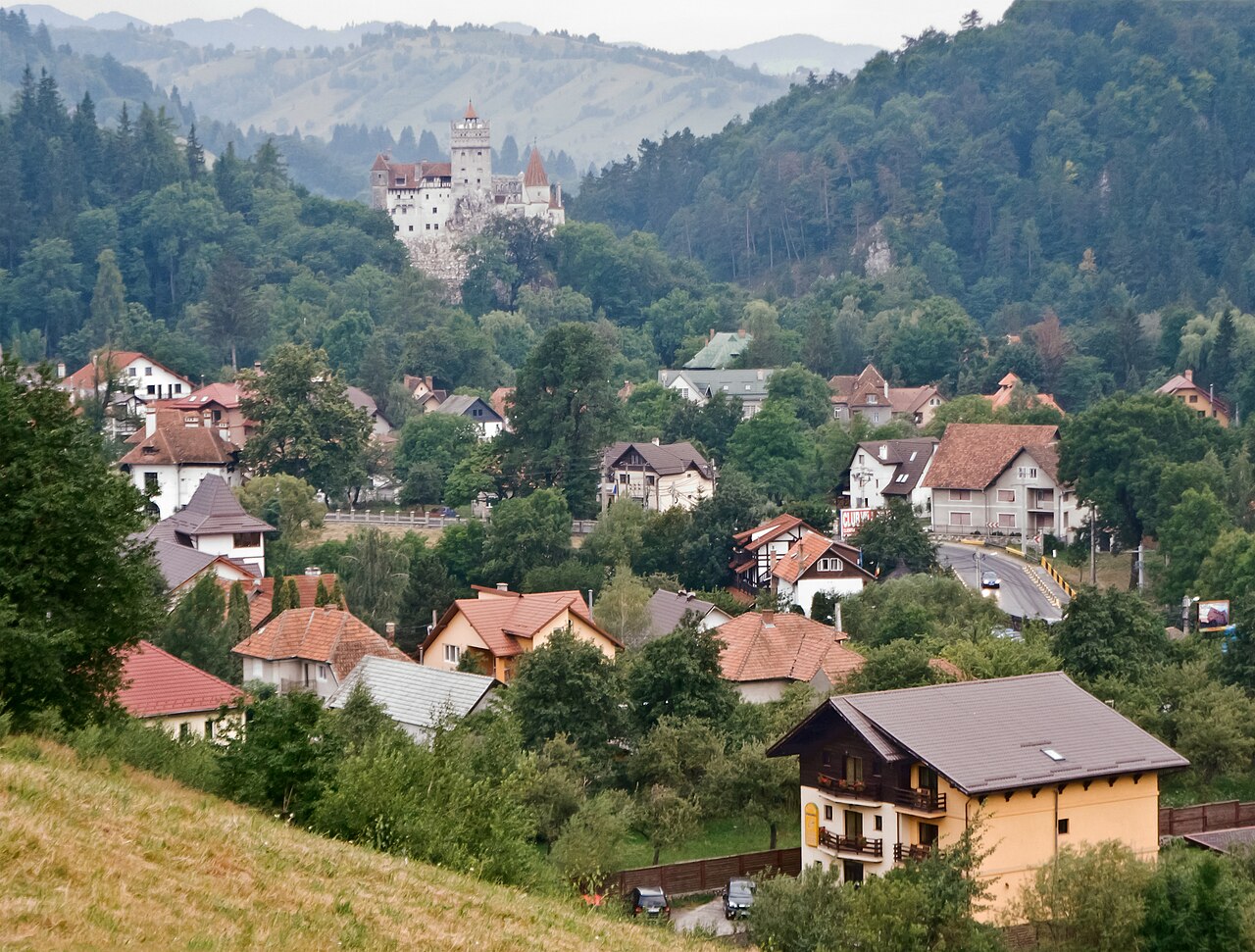 Panorama of Bran village in Romania, red-tiled houses in a valley, with Bran Castle's white towers rising on a forested rock outcrop behind