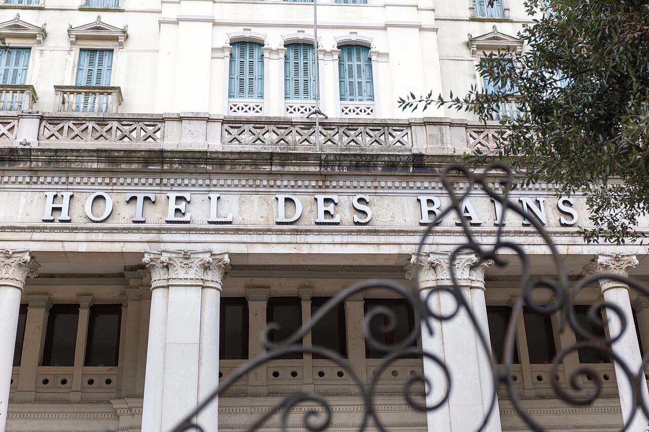 威尼斯丽都岛的Hotel Des Bains建筑正面，古典柱式、装饰性栏杆、蓝色百叶窗，前景铁艺装饰与树木