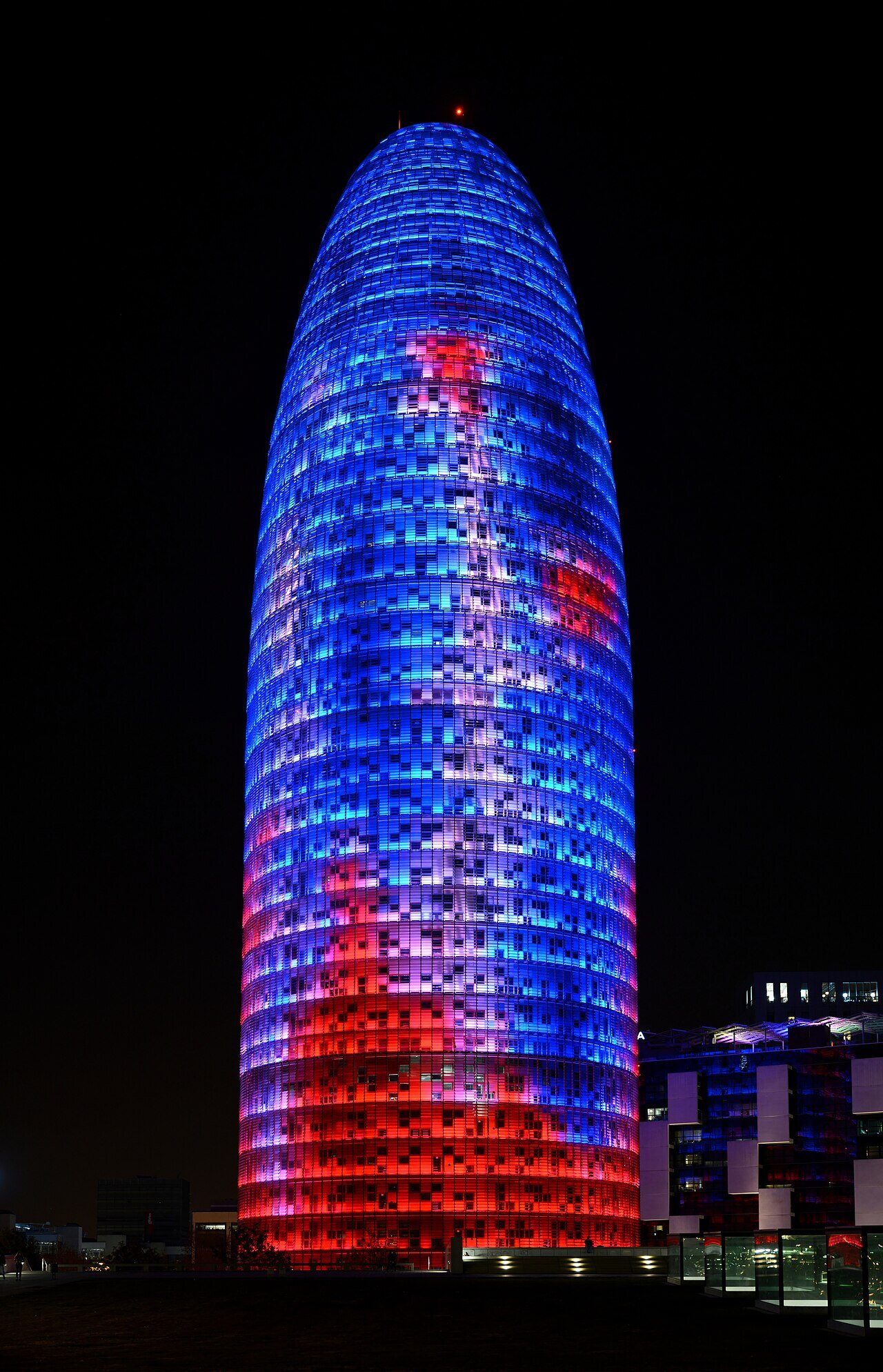 Torre Glòries tower in Barcelona at night, its bullet-shaped facade lit in red and blue LEDs