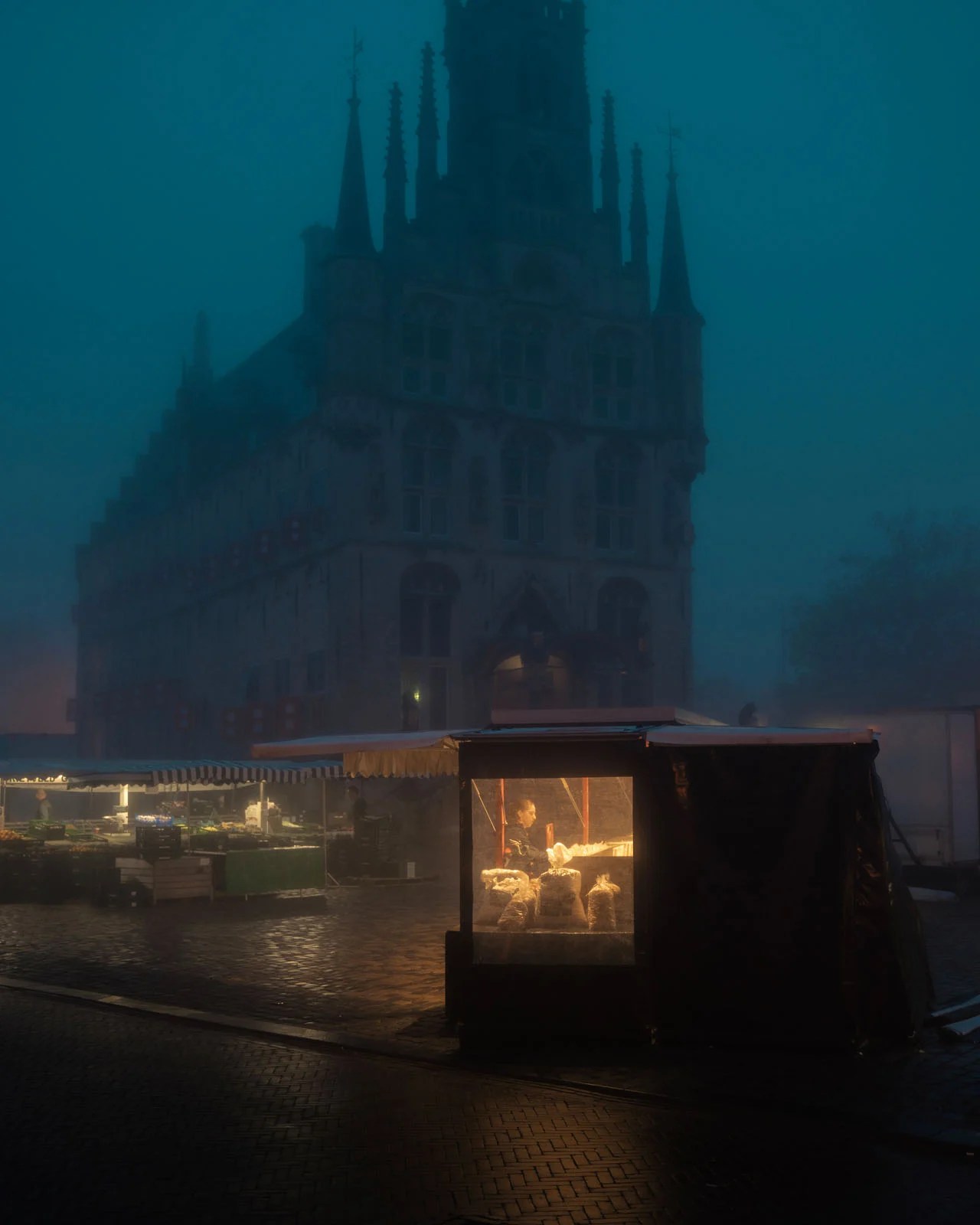 Toplo osvijetljeni tržni &scaron;tand na mokroj kaldrmi ispred gotičkog zdanja u plavoj večernjoj magli, serija Morning Ritual, pobjednik kategorije ulične fotografije