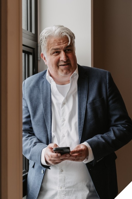 Man in zakelijke kleding staat bij een groot raam in een kantoorruimte en gebruikt zijn telefoon, terwijl zacht daglicht de ruimte verlicht.