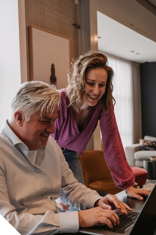 Man en vrouw in zakelijke kleding werken samen aan een laptop aan een tafel in een kantoorachtige omgeving. De man typt terwijl de vrouw over zijn schouder meekijkt; beide hebben een ontspannen en betrokken houding.