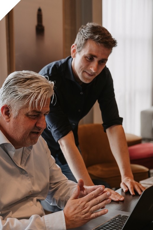 Twee mannen werken samen aan een laptop in een kantooromgeving. De ene man zit aan tafel en maakt een gebaar met zijn handen, terwijl de andere man naast hem staat en meekijkt op het scherm. Op de achtergrond zijn stoelen en zacht daglicht zichtbaar.
