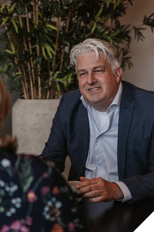 Man in zakelijke kleding zit aan een tafel in een kantooromgeving en houdt een kopje vast tijdens een gesprek. Op de achtergrond zijn een grote plantenbak en zachte binnenverlichting zichtbaar.
