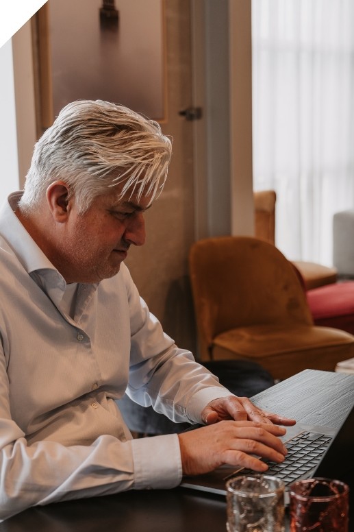Man zit aan een tafel en werkt op een laptop in een rustige kantooromgeving. Op de achtergrond zijn stoelen en zacht daglicht door een raam zichtbaar.