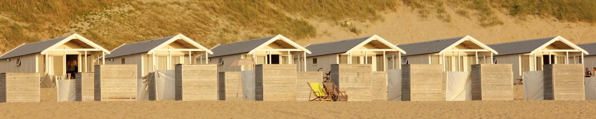 Afbeelding van een rijtje strandhuisjes met 2 ligstoelen ervoor in het zand