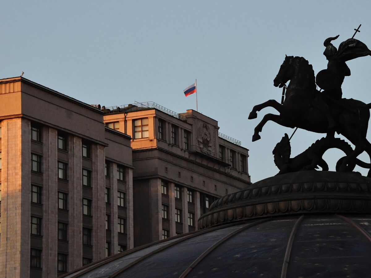 Искусственный интеллект в образе робота сидит за столом, как на допросе, напротив человека в деловом костюме, символизирующего Госдуму. На столе документы и микрофон.