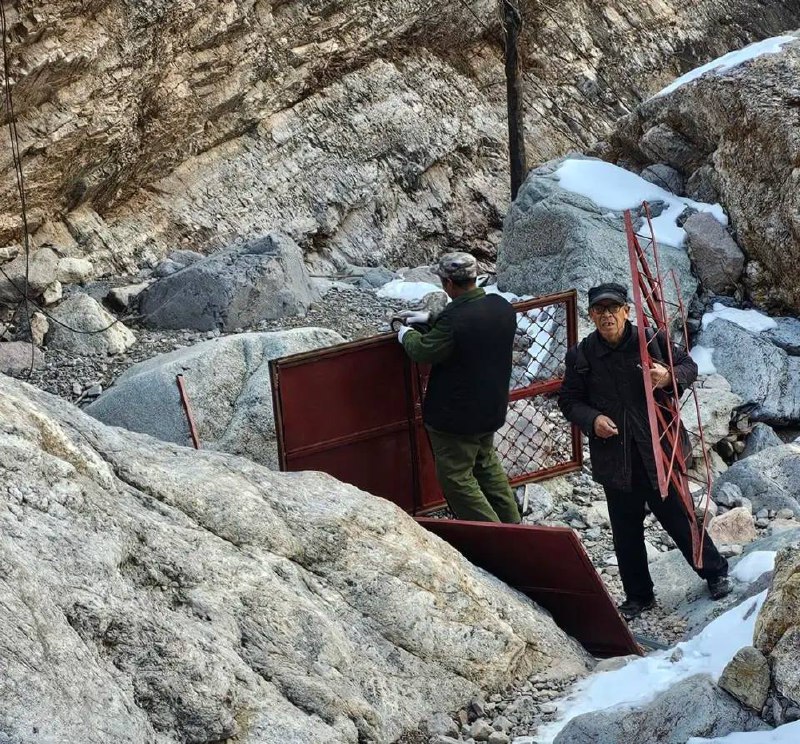 Пожилой китайский мужчина стоит у подножия или на фоне грандиозной, самодельной лестницы, ведущей вверх по склону горы.