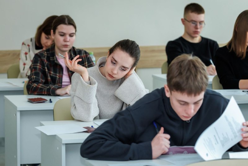 Группа студентов активно участвует в школьной олимпиаде, демонстрируя творческие работы, возможно, под наблюдением жюри или преподавателя, символизируя инновации и личное взаимодействие.