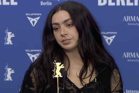 Charli XCX speaking at the Berlinale film festival, with a backdrop related to her album 'Brat'.