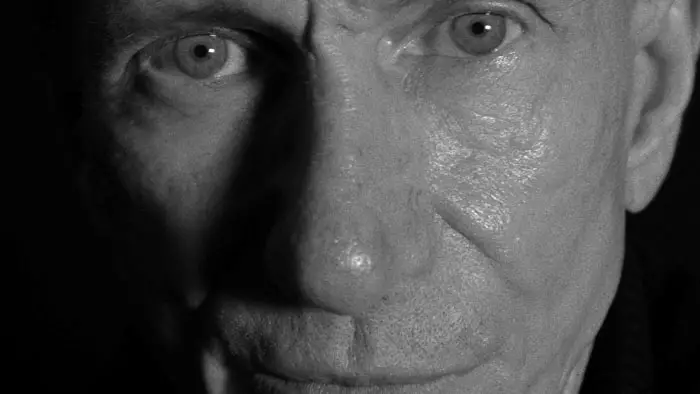 Bill Oberst Jr. in costume as Edgar Allan Poe, holding a microphone, with a dark and intense expression.