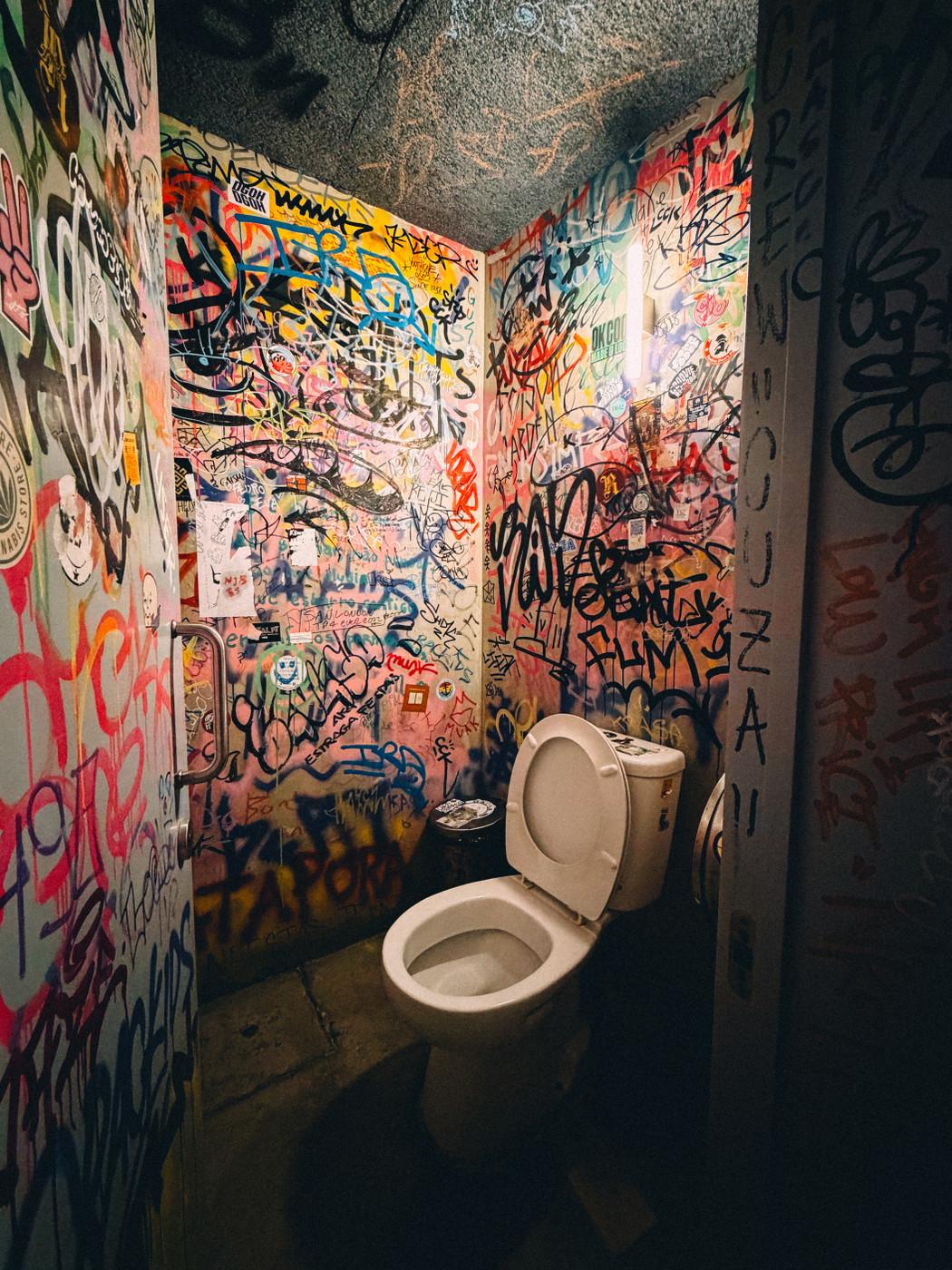 Graffiti-covered public restroom with a white toilet and walls filled with colorful tags, drawings, and stickers.
