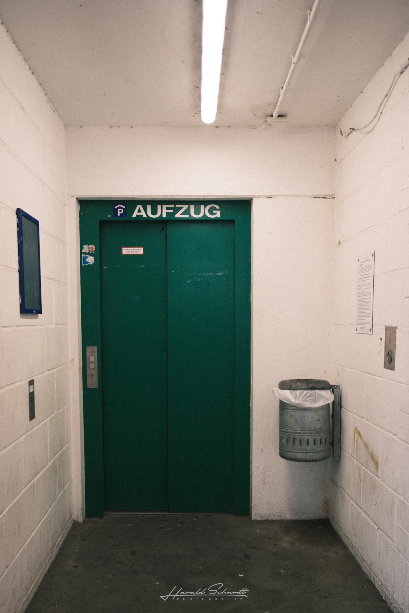 A green elevator inside a concrete parking garage. White walls, fluorescent lights, and a trash bin beside the doors.