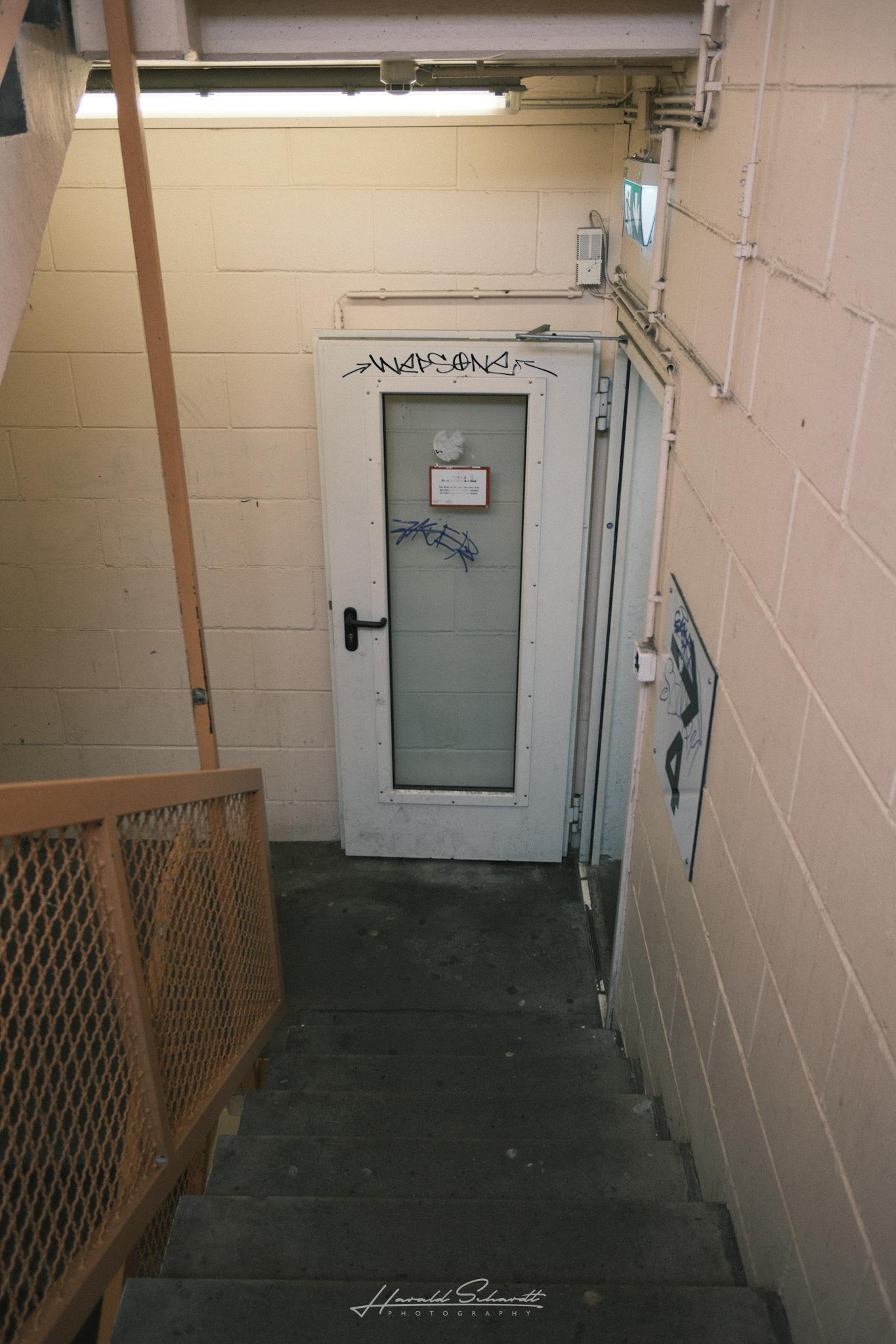 A narrow concrete stairwell inside the parking garage leading down to a gray metal door with a few graffiti tags.
