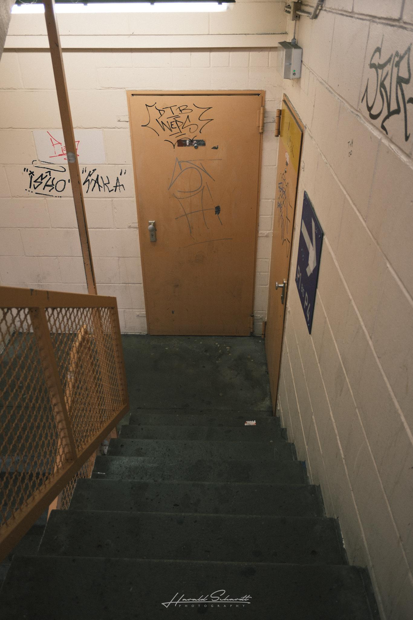 A second stairwell inside the parking garage leading down to a wooden door covered with graffiti.