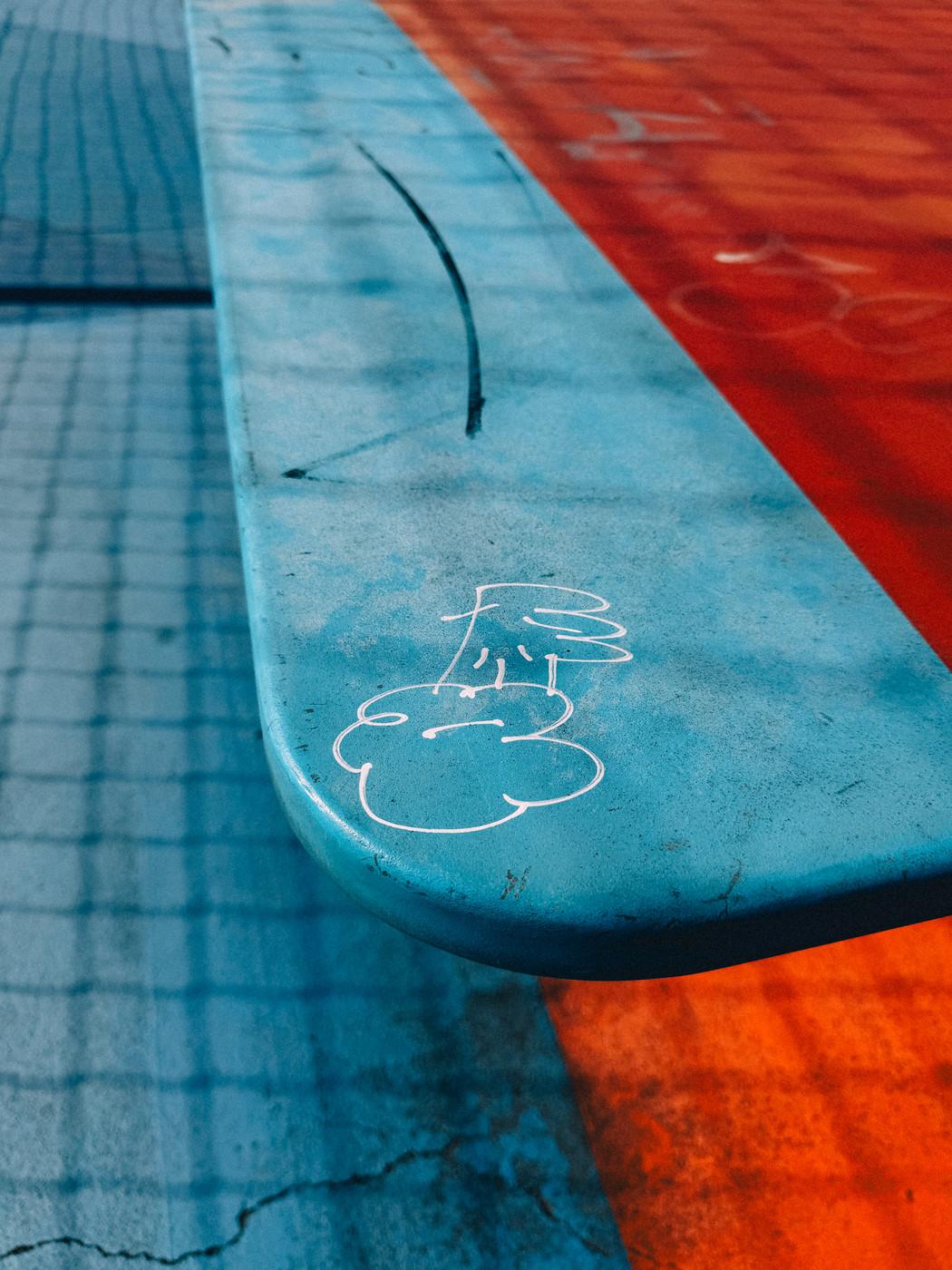 Close-up of a weathered blue table tennis table with a simple white graffiti drawing, set against a background of red and blue pavement.