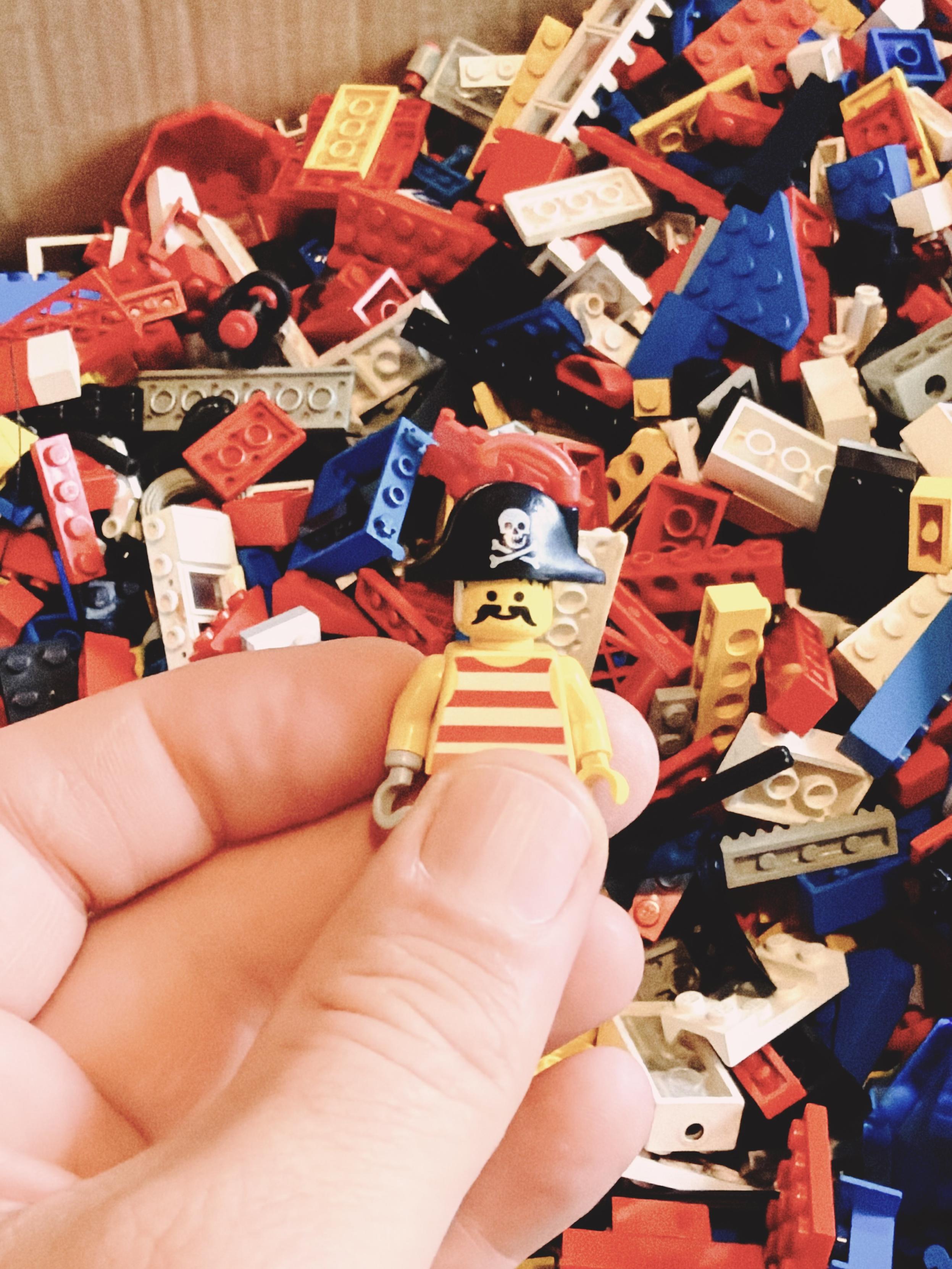 A classic LEGO pirate minifigure with a striped shirt, mustache and black pirate hat is held in front of a box full of vintage LEGO bricks.