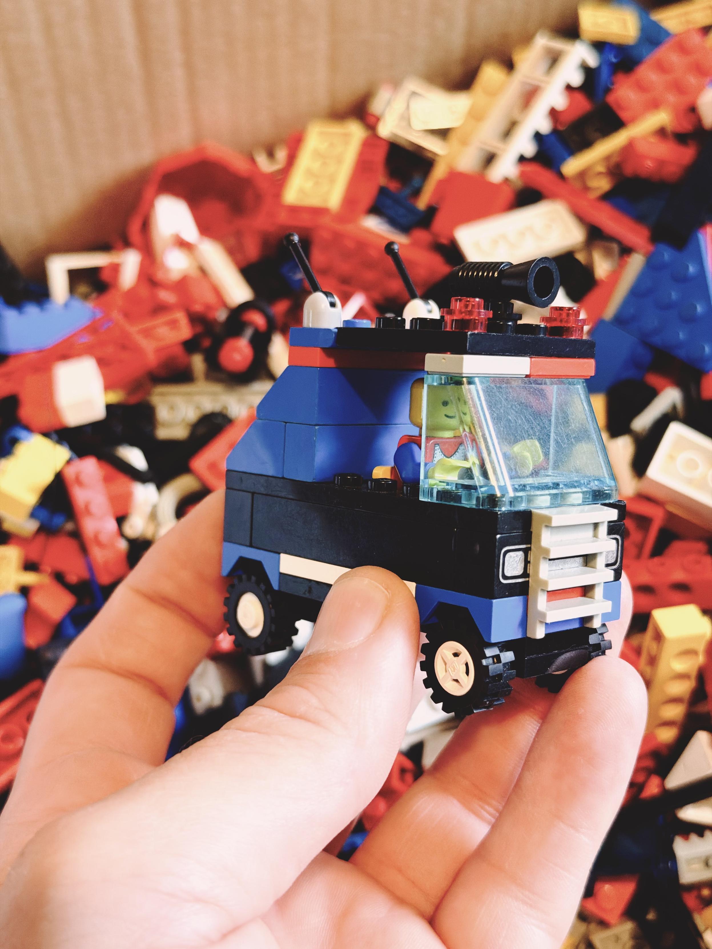 A small blue LEGO police van with a smiling minifigure driver is held in front of a box of old LEGO bricks.