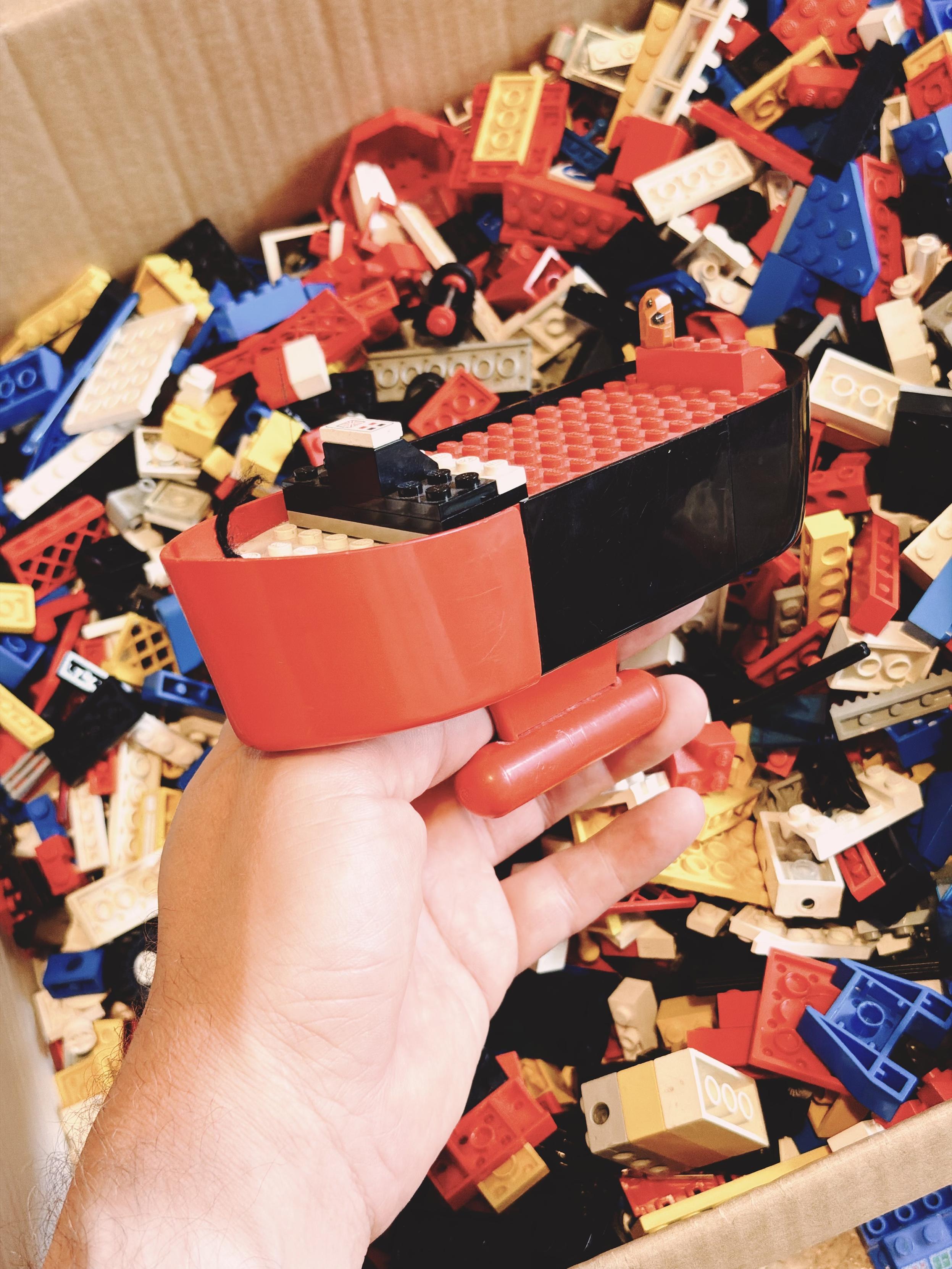 A classic red and black LEGO boat hull from the late 1970s is held above a box filled with vintage LEGO bricks.