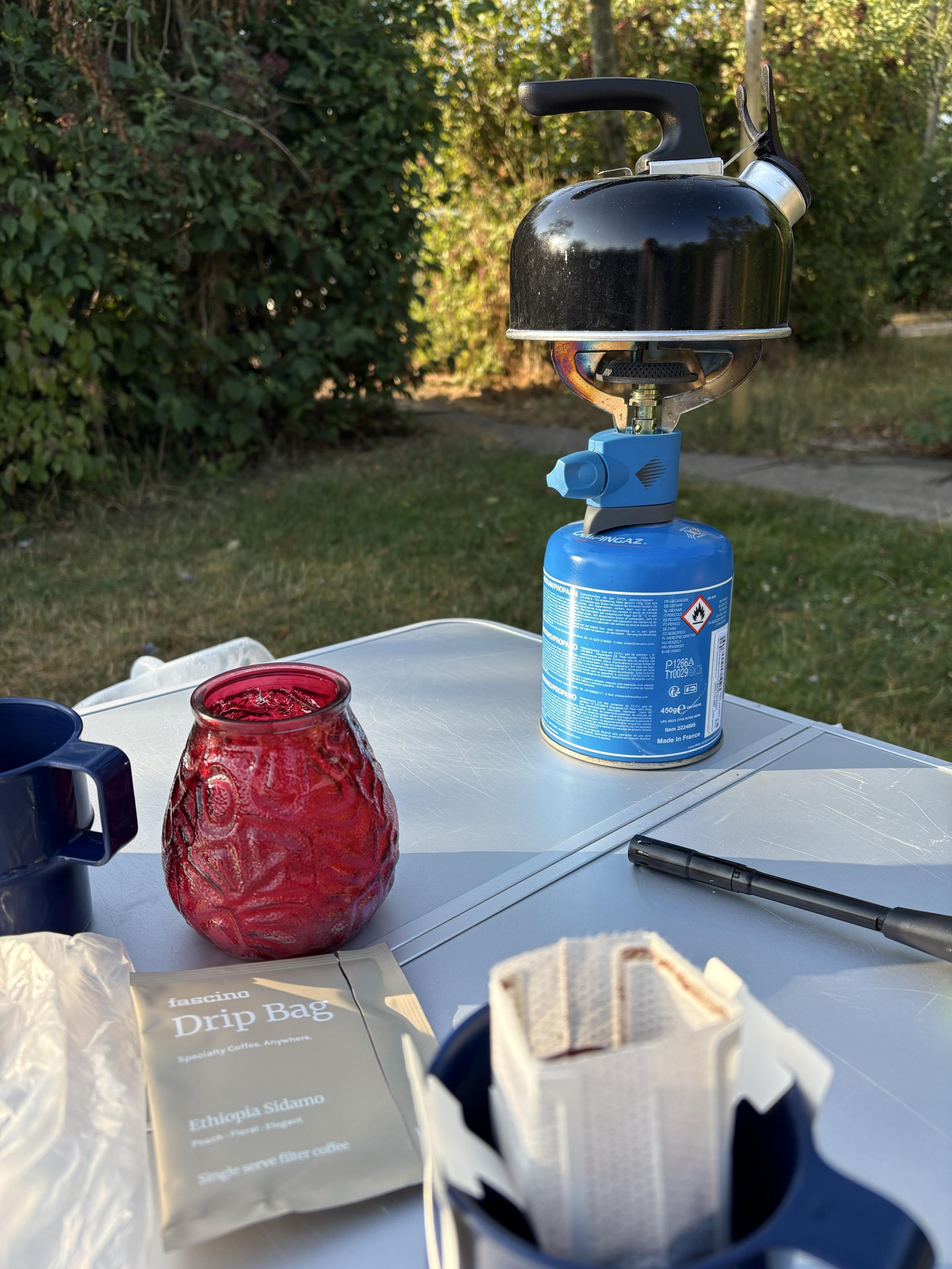 Camping table with a coffee cup with a single serve drip filter with coffee in it hanging in the cup and a little kettle boiling water on a camping gas behind it