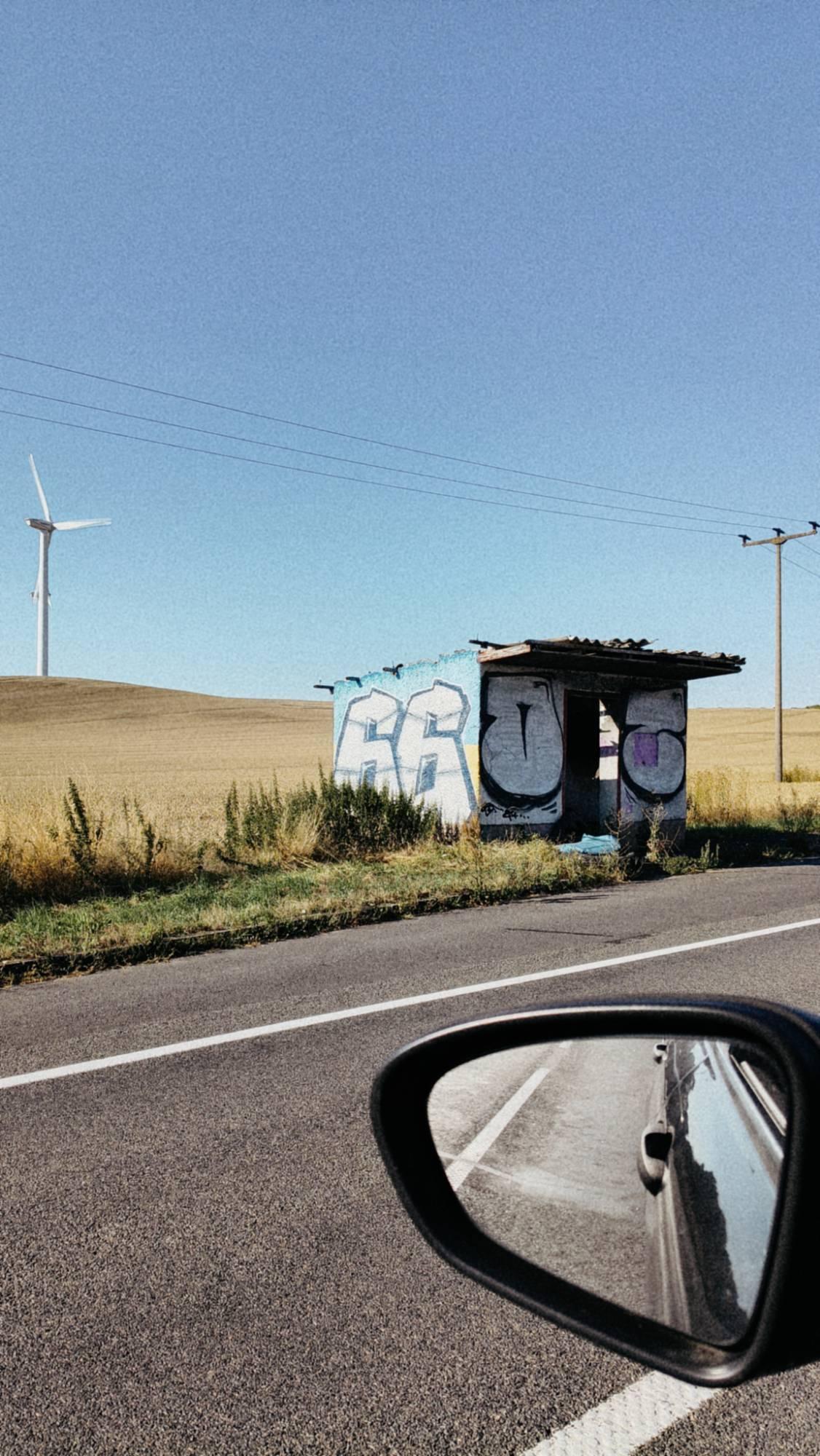 An old bus stop and a field near a road. Car mirror in front.