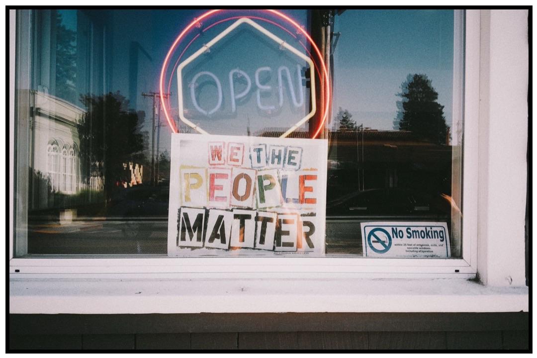 A shop window features a bright neon "OPEN" sign and a colorful poster that reads "WE THE PEOPLE MATTER." A "No Smoking" sign is also visible.