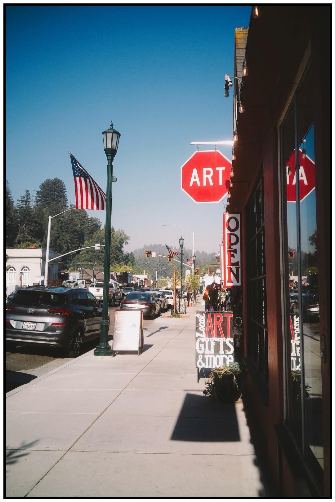 A sunny street scene featuring a sidewalk lined with parked cars, storefronts, and decorative signs. A notable red stop sign indicates "ART." An American flag is displayed, and pedestrians can be seen in the distance.