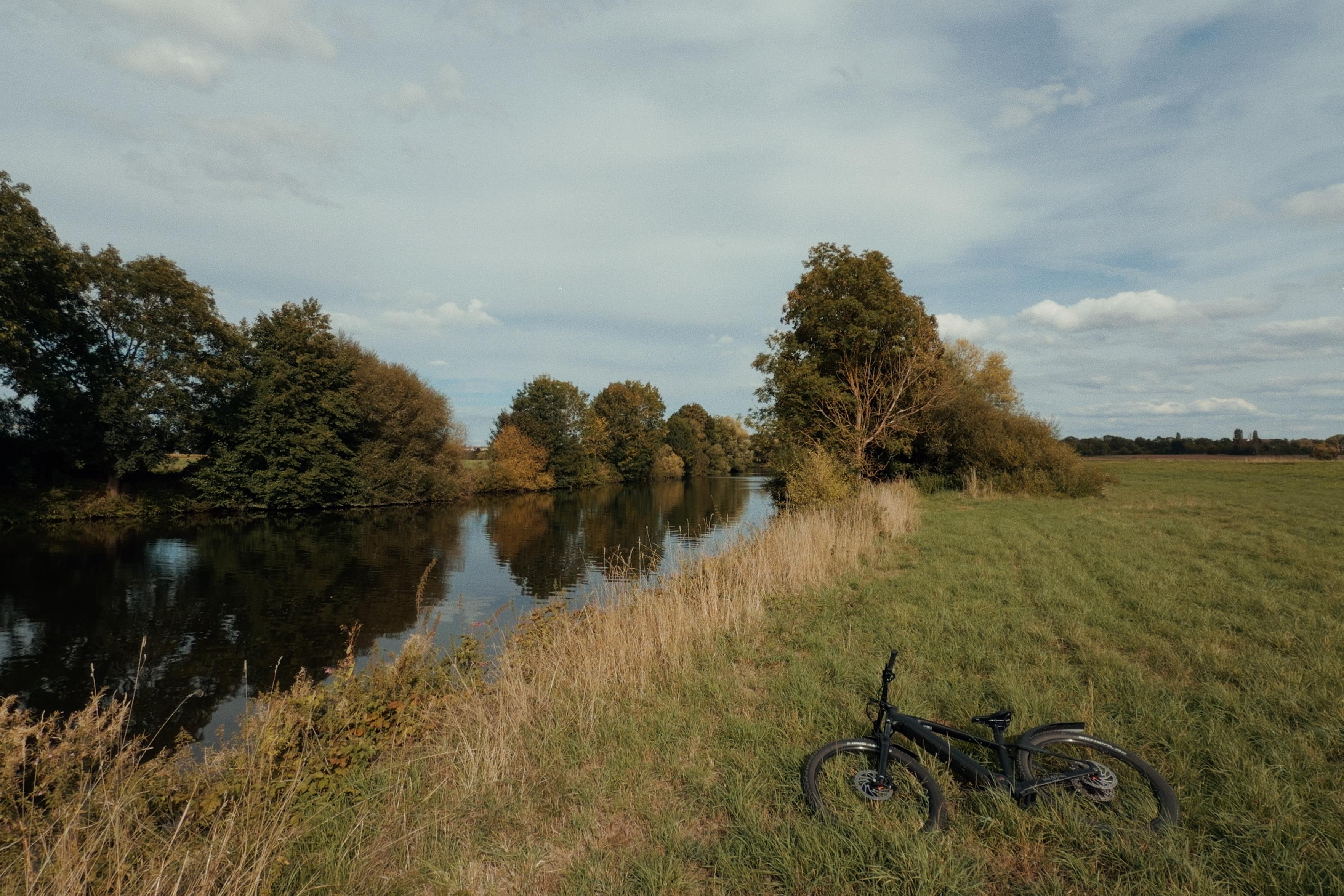 A black bicycle lies in tall grass beside a calm river, surrounded by trees and open fields under a partly cloudy blue sky.