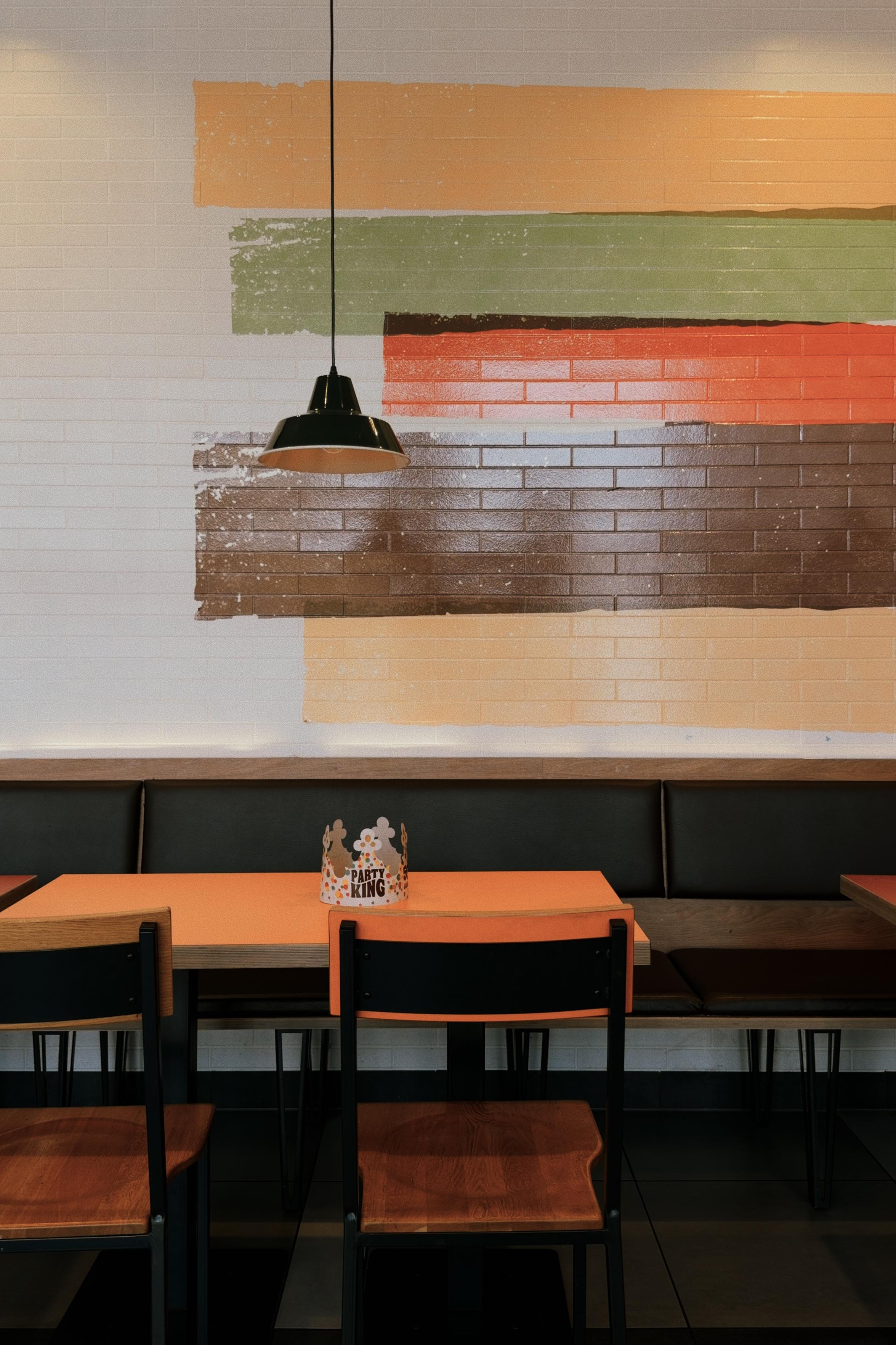 Empty restaurant seating area with orange tables, wooden chairs and a black bench. A small “Party King” paper crown sits on the table beneath a hanging lamp, with broad painted color stripes on the brick wall behind it.