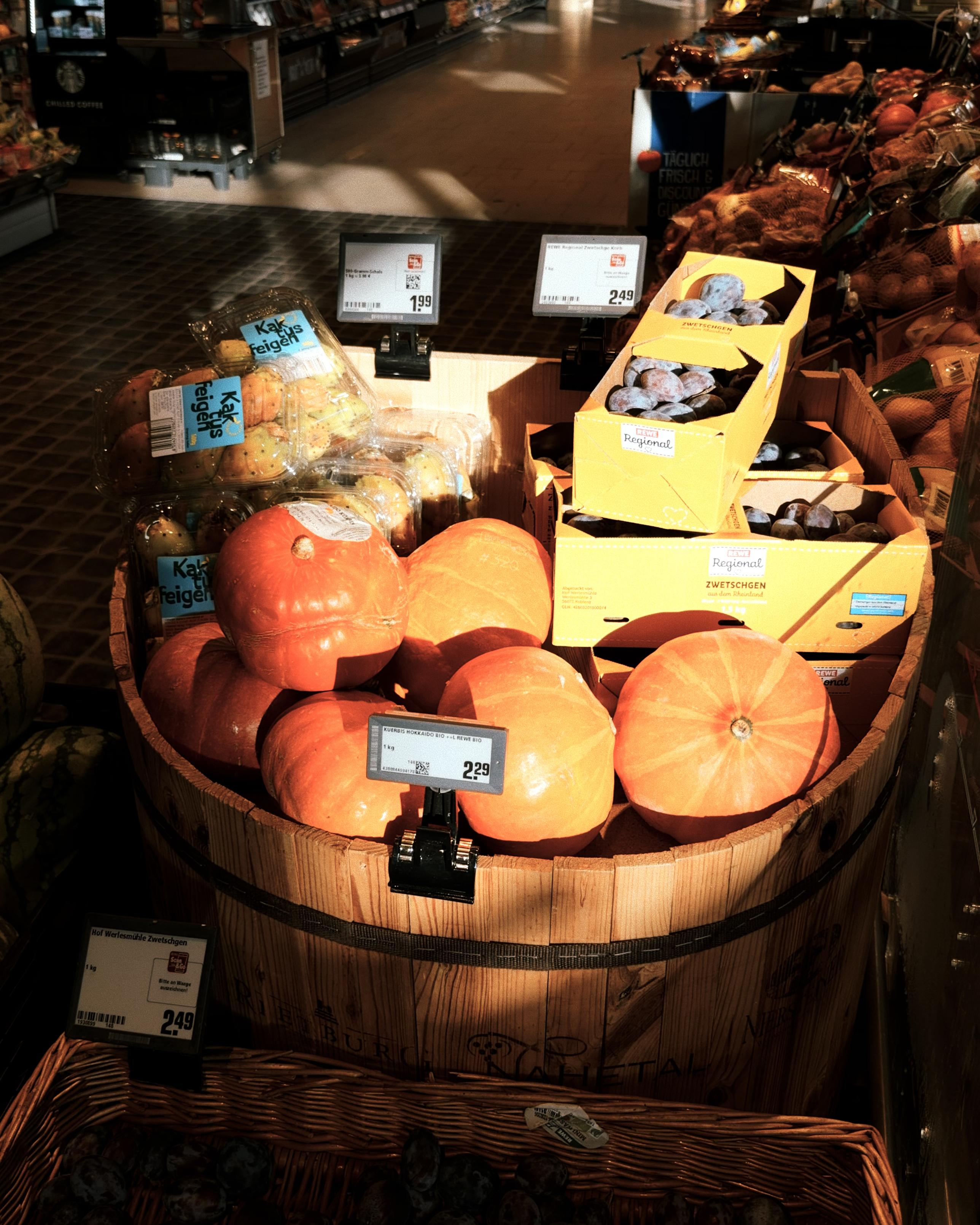Warm evening sunlight falls across a supermarket display of bright orange pumpkins, with boxes of plums and figs behind them.