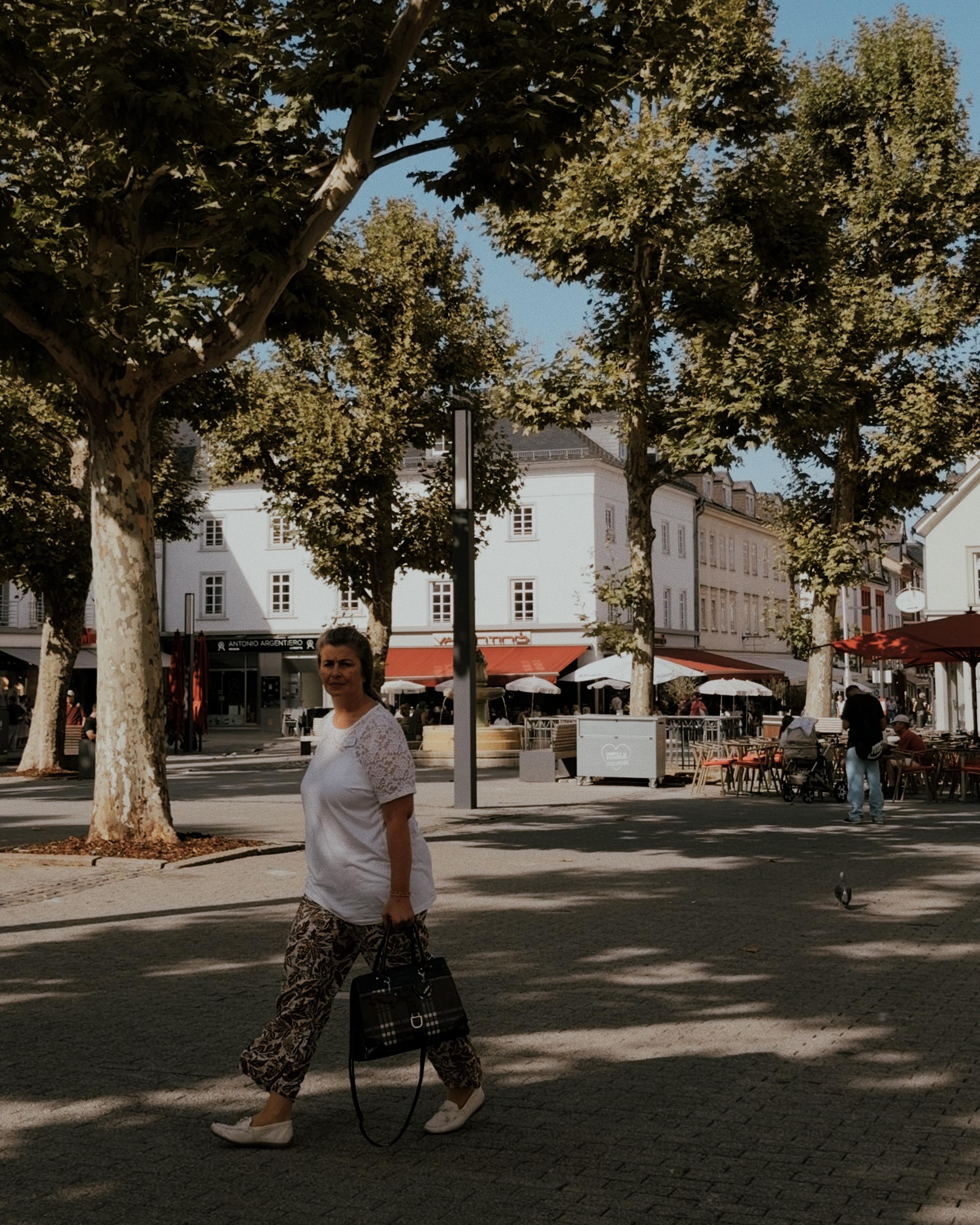 Woman walking across a sunlit town square beneath large leafy trees, with cafés and old buildings in the background.