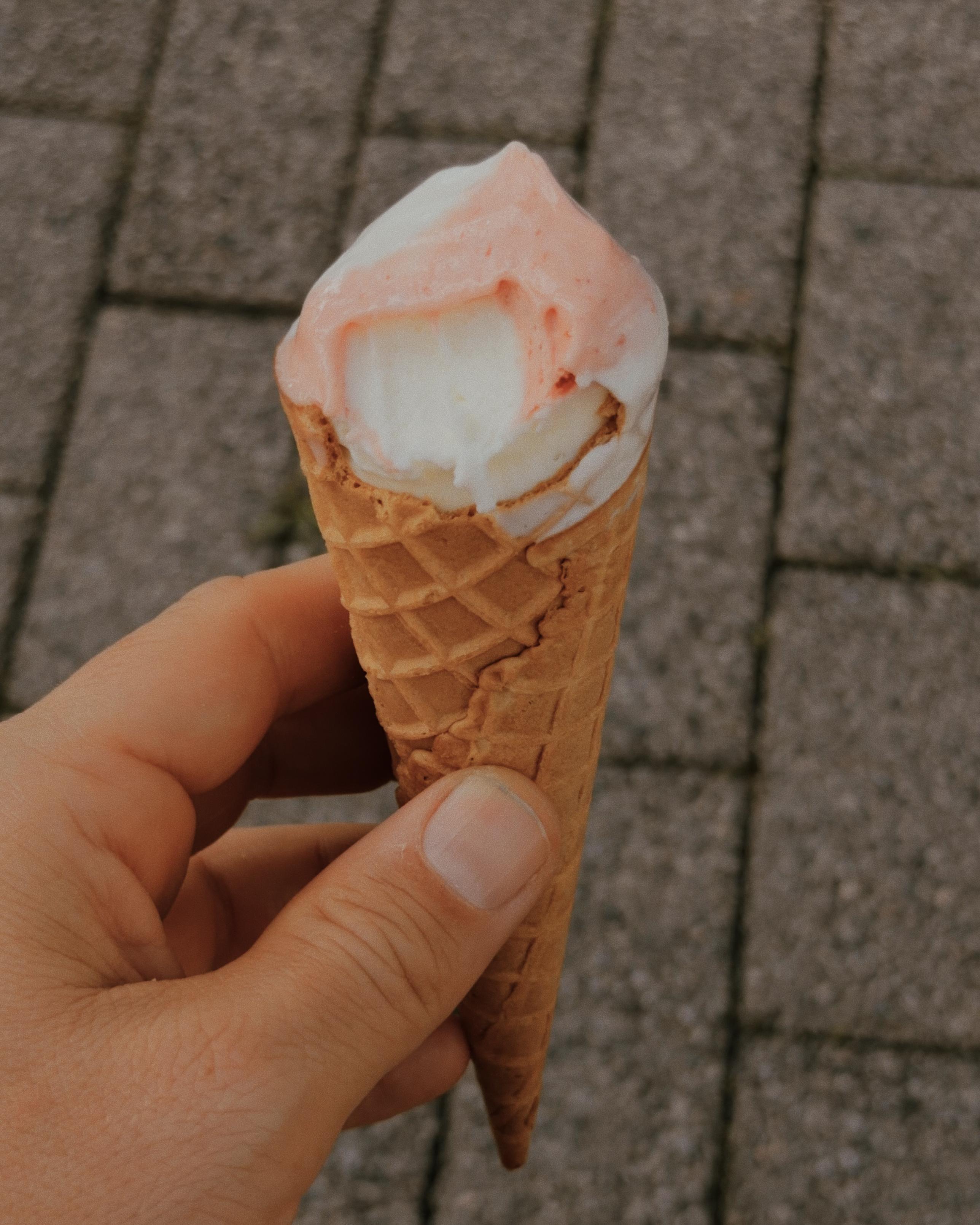 Hand holding a partly eaten pink-and-white ice cream cone over a paved street.