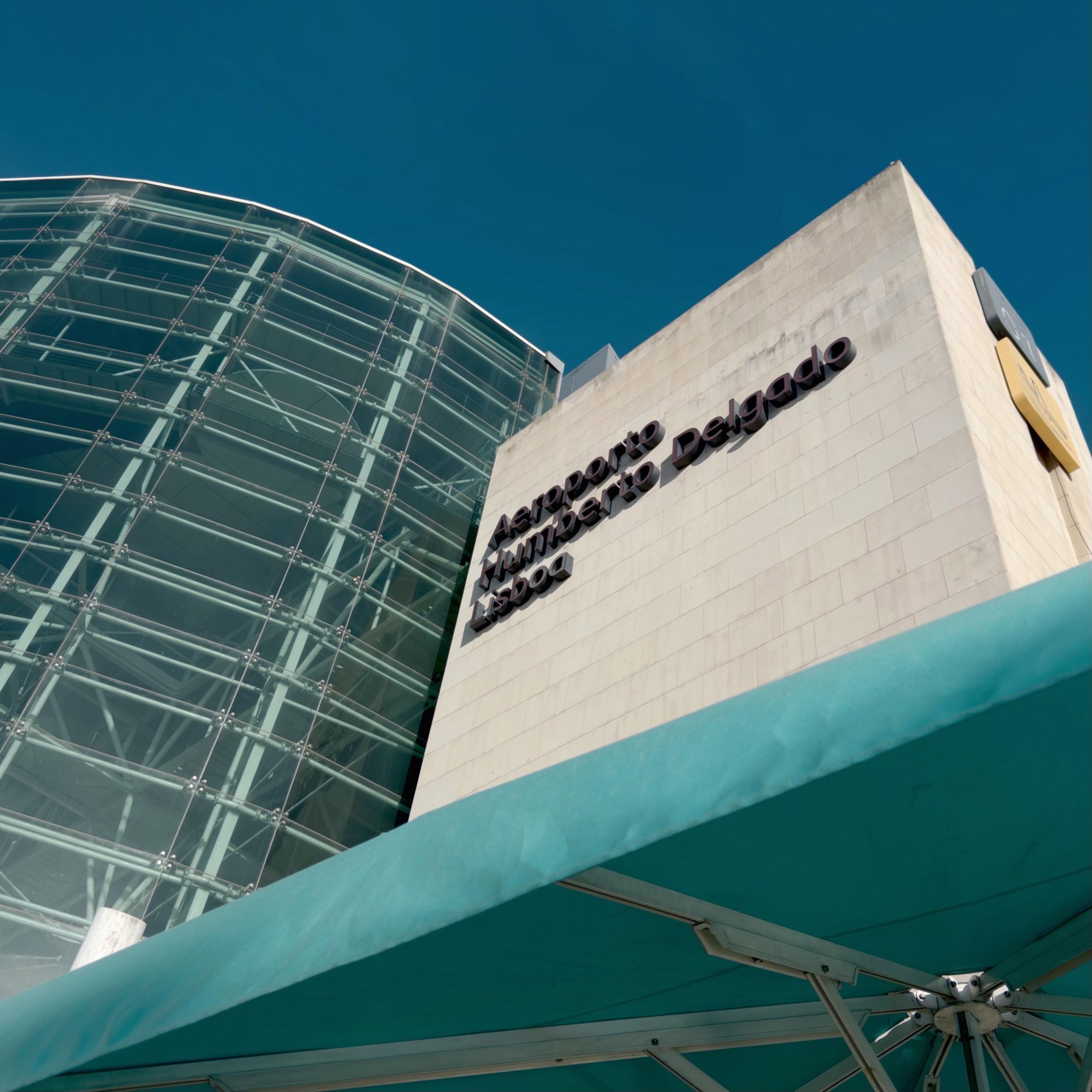 Modern white building with "Aeropuerto Umberto Delgado Lisboa" text on facade, glass curved structure to left, turquoise patio umbrella visible in foreground, clear blue sky above.