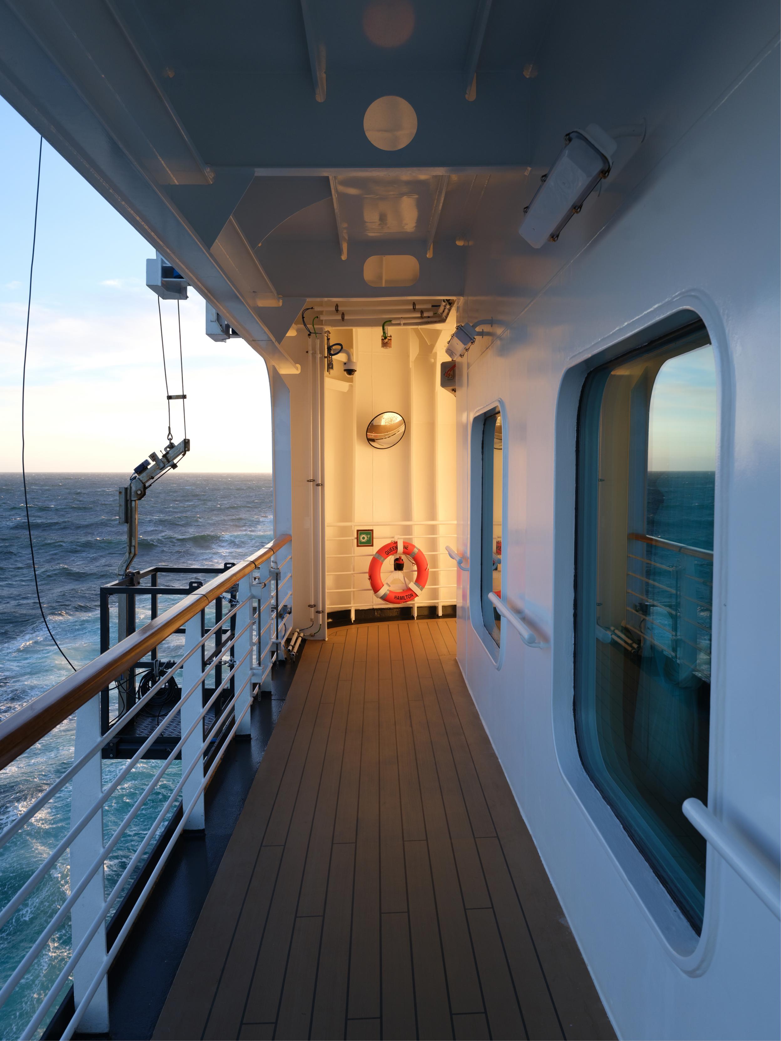 A boat's interior with a wooden floor, a white wall, a red life-saving ring, and a window overlooking the ocean.