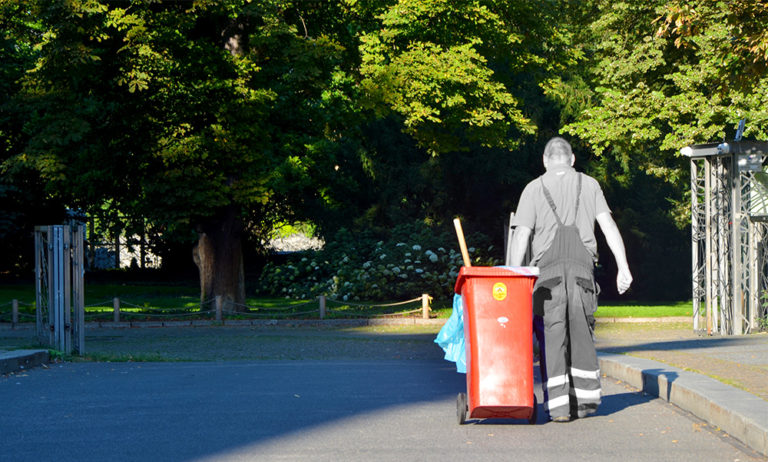 A city street cleaner seen from behind, walking away while pulling a large wheeled trash bin.